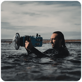 Man standing waist-deep in the ocean holding a blue underwater camera housing with a dome lens port, preparing to shoot in moody natural light. The setup highlights professional gear used for surf and ocean photography.